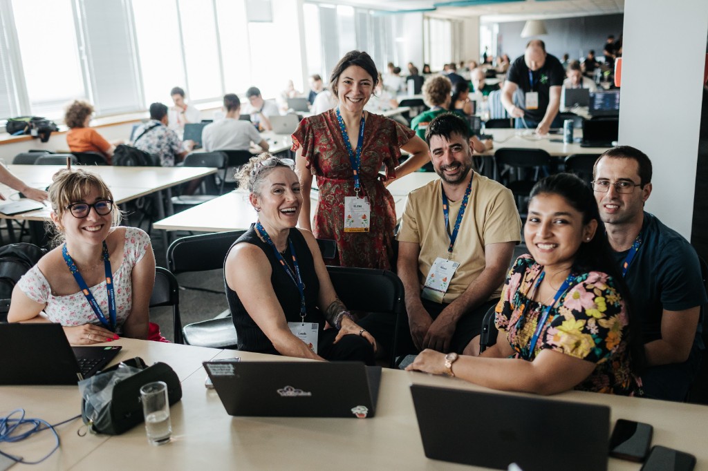 Contributors smile around laptops during a conference sprint, with more people working at tables in a bright room behind them.